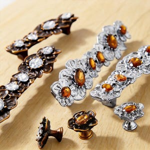May include: Decorative cabinet hardware in various styles. The hardware includes bronze and silver-toned pulls and knobs, each adorned with clear and amber-colored crystal-like accents. The floral designs add a touch of elegance.