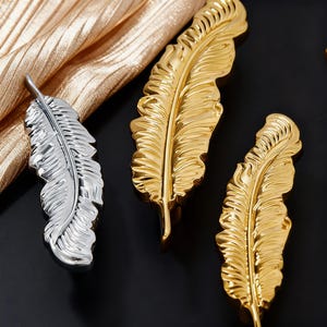 May include: Three decorative feather-shaped objects. Two are gold-colored, and one is silver. The feathers have detailed veining and are arranged on a black surface with a gold fabric in the background. The feathers are likely decorative accents.