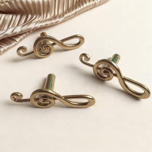 Può includere: Tre pomelli dorati per mobili a forma di chiave di violino. I pomelli hanno una finitura metallica liscia e un piccolo accento verde sullo stelo. Sono disposti su una superficie bianca con tessuto dorato sullo sfondo.