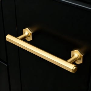 Op de afbeelding: Een goudkleurige kastgreep met een geometrisch ontwerp. De handgreep is een lange, rechte staaf met zeshoekige uiteinden. De greep is gemonteerd op een zwarte kastdeur.