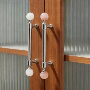 May include: Two cabinet door pulls with a polished chrome finish. Each pull has a long, straight handle and a decorative sphere at each end. One pull has white spheres, the other has pink spheres. The pulls are mounted on a wooden cabinet.