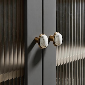 Pode incluir: Close-up de dois puxadores de porta de armário. Os puxadores têm um centro de mármore branco com veios pretos, rodeado por um anel de metal dourado. Os puxadores são montados numa porta de armário cinza escuro. O fundo tem um design canelado vertical.