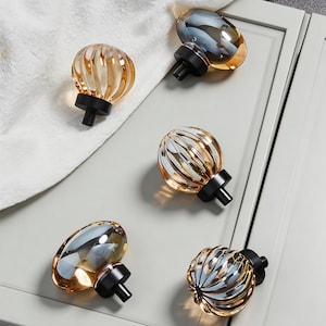 May include: Five decorative cabinet knobs with a clear, amber-toned glass design and ribbed texture. Each knob has a black base and stem. The knobs are arranged on a light grey surface. The knobs are suitable for cabinets.