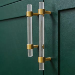 Peut inclure: Poignées de placard en acrylique transparent avec des accents métalliques dorés montées sur une porte d'armoire vert foncé. Les poignées cylindriques ont un design moderne, ajoutant une touche d'élégance à l'armoire.