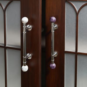 以下が含まれることがあります： 磨かれたクロム仕上げのキャビネットドアハンドルが2つ。片方のハンドルは白い大理石の球体、もう片方は紫色の球体です。ドアは濃い茶色で、すりガラスのパネルが付いています。ハンドルは約30cmです。
