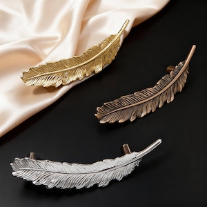 Può includere: Tre pomelli decorativi a forma di piuma, in oro, bronzo e argento. I pomelli sono in metallo e presentano un design dettagliato a piuma. Il pomello dorato è su un tessuto di raso color crema, gli altri su sfondo nero.