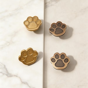 Puede incluir: Cuatro tiradores de armario con forma de pata en acabado dorado y bronce. Los tiradores están dispuestos en un patrón cuadrado sobre un fondo de mármol blanco. Cada tirador presenta un diseño detallado de huella de pata, ideal para mascotas.