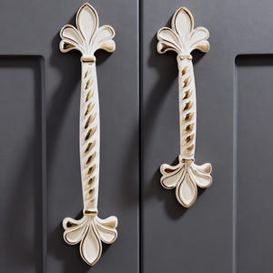 Op de afbeelding: Twee sierlijke kastgrepen met een witte en gouden afwerking. De grepen hebben aan elk uiteinde een fleur-de-lis-ontwerp en een gedraaide, geribbelde midden. Ze zijn gemonteerd op een grijze kastdeur.