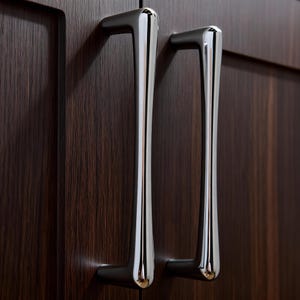 Könnte beinhalten: Nahaufnahme von zwei polierten Chrom-Schrankgriffen vor einer dunklen Holzmaserungsoberfläche. Die Griffe haben ein schlankes, modernes Design mit einer geschwungenen Form und einer reflektierenden Oberfläche, die der Schrankwand Eleganz verleiht.