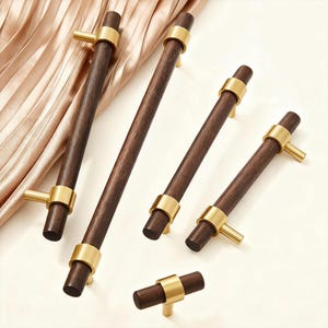 Può includere: Una collezione di maniglie per mobili in legno con dettagli in ottone. Le maniglie hanno un'impugnatura in legno cilindrica marrone scuro, un anello color oro e una maniglia in ottone. Le maniglie sono di dimensioni variabili e sono disposte su uno sfondo chiaro.