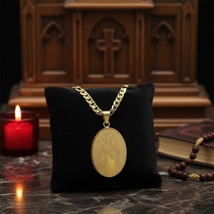 Peut inclure: Un collier pendentif doré avec un pendentif ovale représentant une figure religieuse. Le collier a une chaîne épaisse et est présenté sur un coussin de velours noir. Une bougie rouge allumée et une croix en bois sont en arrière-plan.