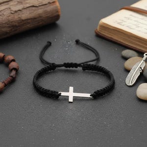 Peut inclure: Un bracelet tressé noir avec un charm en forme de croix argentée. Le bracelet est réglable et posé sur une surface sombre. D'autres accessoires, dont un pendentif en forme de plume et un bracelet de perles en bois, sont également visibles.