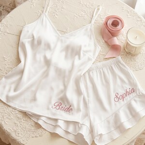 Könnte beinhalten: Weißes Satin-Pyjama-Set mit einem Camisole-Top und Shorts. Das Wort "Bride" ist auf dem Oberteil und der Name "Sophia" auf den Shorts in rosa Faden gestickt. Ein rosa Band und eine Kerze befinden sich ebenfalls auf dem Tisch.