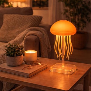 Può includere: Lampada arancione a forma di medusa con base trasparente e tentacoli fluenti su un tavolo di legno. La lampada emette una calda luce, illuminando l'ambiente circostante. Una candela, una pianta e dei libri sono anche sul tavolo.