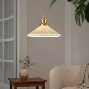 May include: A retro pendant light with a ribbed, cream-coloured glass shade and brass fittings. The light is suspended by a gold-coloured cord. A small wooden table holds a vase with dried flowers, and a cream-coloured chair is in the background.
