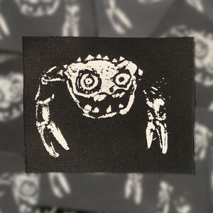 May include: A rectangular black fabric patch featuring a white screen-printed graphic of a stylized crab with large circular eyes, a textured shell, and two prominent claws. The design has a distressed, ink-stamped appearance on the black textile.