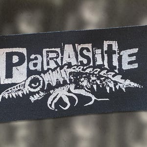 May include: Black rectangular patch featuring the word "PARASITE" in white, block letters. Beneath the word is a white illustration of a creature with sharp teeth and tentacles. The patch is designed for clothing or accessories.