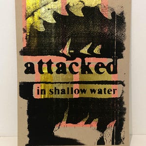 May include: A rectangular art print on a tan background. The print features the words "attacked" and "in shallow water" in black text. The design includes black, gold, and coral-colored abstract shapes.