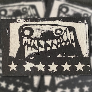May include: A black and white patch with a monster face design. The image shows a creature with sharp teeth and large eyes on a white background, framed by a black border. White stars are along the bottom edge. Suitable for clothing or accessories.