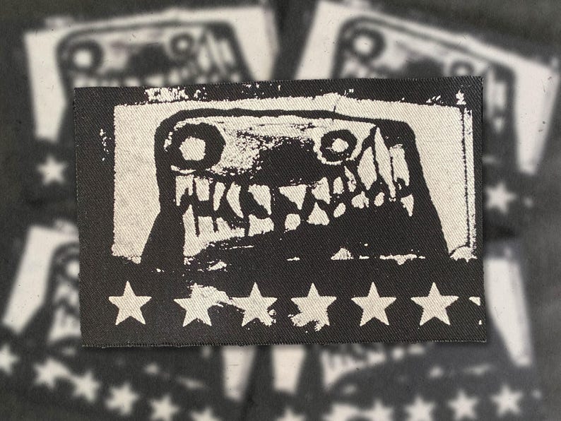 Puede incluir: Parche en blanco y negro con un dise&ntilde;o de cara de monstruo. La imagen muestra una criatura con dientes afilados y ojos grandes sobre un fondo blanco, enmarcada por un borde negro. Estrellas blancas en el borde inferior. Para ropa o accesorios.
