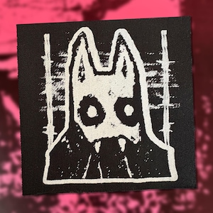 Könnte beinhalten: Ein quadratischer schwarzer Stoffaufnäher mit einer weißen Siebdruckgrafik einer stilisierten Kreatur mit spitzen Ohren, großen kreisförmigen Augen und sichtbaren Fangzähnen. Das Design ist von vertikalen Stacheldrahtmustern eingerahmt.