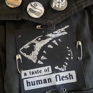 Pode incluir: Tecido preto com três botões redondos e um patch retangular. Os botões exibem as palavras "SURF", "Attacked in shallow water" e um gráfico. O patch diz "a taste of human flesh" e é preso com alfinetes de segurança.