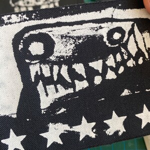 Puede incluir: Parche de tela en blanco y negro con un dise&ntilde;o de cara de monstruo y la palabra "WKSW". El borde inferior tiene formas de estrellas blancas. El parche est&aacute; sujeto por una mano.