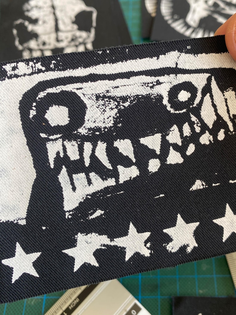 Puede incluir: Parche de tela en blanco y negro con un dise&ntilde;o de cara de monstruo y la palabra "WKSW". El borde inferior tiene formas de estrellas blancas. El parche est&aacute; sujeto por una mano.