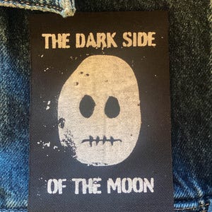 Puede incluir: Un parche rectangular negro con el texto "THE DARK SIDE OF THE MOON" en blanco. El parche presenta un gráfico de calavera blanca con dos agujeros para los ojos y una boca cosida. El parche está cosido a una chaqueta vaquera.