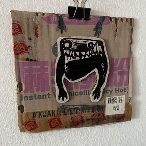 May include: A piece of cardboard with a black monster sticker attached with a binder clip. The cardboard has faded pink and red text, with the words "Instant Vermicelli Spicy Hot" visible. A small white label reads "105g*20".