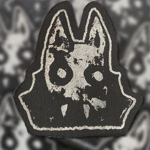 Könnte beinhalten: Schwarzer Aufnäher mit einer silbernen, stilisierten Illustration eines Hundegesichts. Der Aufnäher hat ein strukturiertes Aussehen und einen weißen Rand. Der Hintergrund zeigt ein verschwommenes Muster ähnlicher Hundegesicht-Illustrationen.