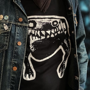 Könnte beinhalten: Ein schwarzes T-Shirt mit einer weißen Grafik eines Wesens mit scharfen Zähnen und Krallen, getragen unter einer Jeansjacke. Die Jacke hat kupferfarbene Knöpfe und dekorative Anstecknadeln. Ein schwarzes Halstuch ist am Hals zu sehen.