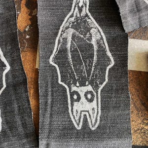 Könnte beinhalten: Mehrere graue Denim-Patches mit einem weißen Fledermaus-Design. Die Fledermaus ist in einer stilisierten, symmetrischen Illustration dargestellt, mit ausgebreiteten Flügeln und nach unten gerichtetem Kopf. Die Patches sind wahrscheinlich für Kleidung.