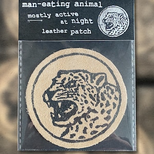 May include: A circular tan leather patch featuring a black jaguar illustration. The patch is packaged in clear plastic. The packaging above the patch reads "man-eating animal", "mostly active at night", and "leather patch".