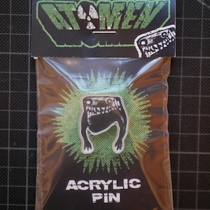 Könnte beinhalten: Schwarzer und weißer Acryl-Pin mit einem Cartoon-Monster-Design. Der Pin ist auf einer schwarzen Rückkarte mit dem Text "ACRYLIC PIN" und dem Markennamen "ATOMEV" in Grün verpackt. Der Pin befindet sich in einer transparenten Plastiktüte.