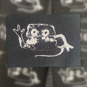 Puede incluir: Un parche rectangular de tela negra con un gráfico serigrafiado en blanco de una criatura estilizada con grandes ojos circulares, una boca ancha y extremidades largas. La criatura está representada con un estilo de tinta texturizada.