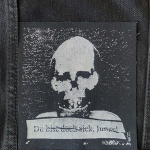 May include: Black and white skull patch with the text "Du bist doch sick, Junge!" sewn onto a black denim jacket. The skull is a stark, high-contrast image, and the text is in a simple sans-serif font.