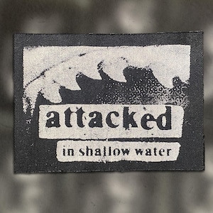 May include: A rectangular black patch with white text and a wave graphic. The text reads "attacked" above "in shallow water". The patch has a textured appearance, suggesting it is made of fabric.