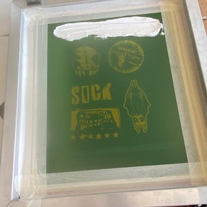 Puede incluir: Un marco de serigraf&iacute;a con un fondo verde que presenta dise&ntilde;os gr&aacute;ficos amarillos, incluyendo una calavera, un murci&eacute;lago y la palabra "SOCK". Tinta blanca se aplica en la parte superior del marco. El marco est&aacute; asegurado con abrazaderas met&aacute;licas.