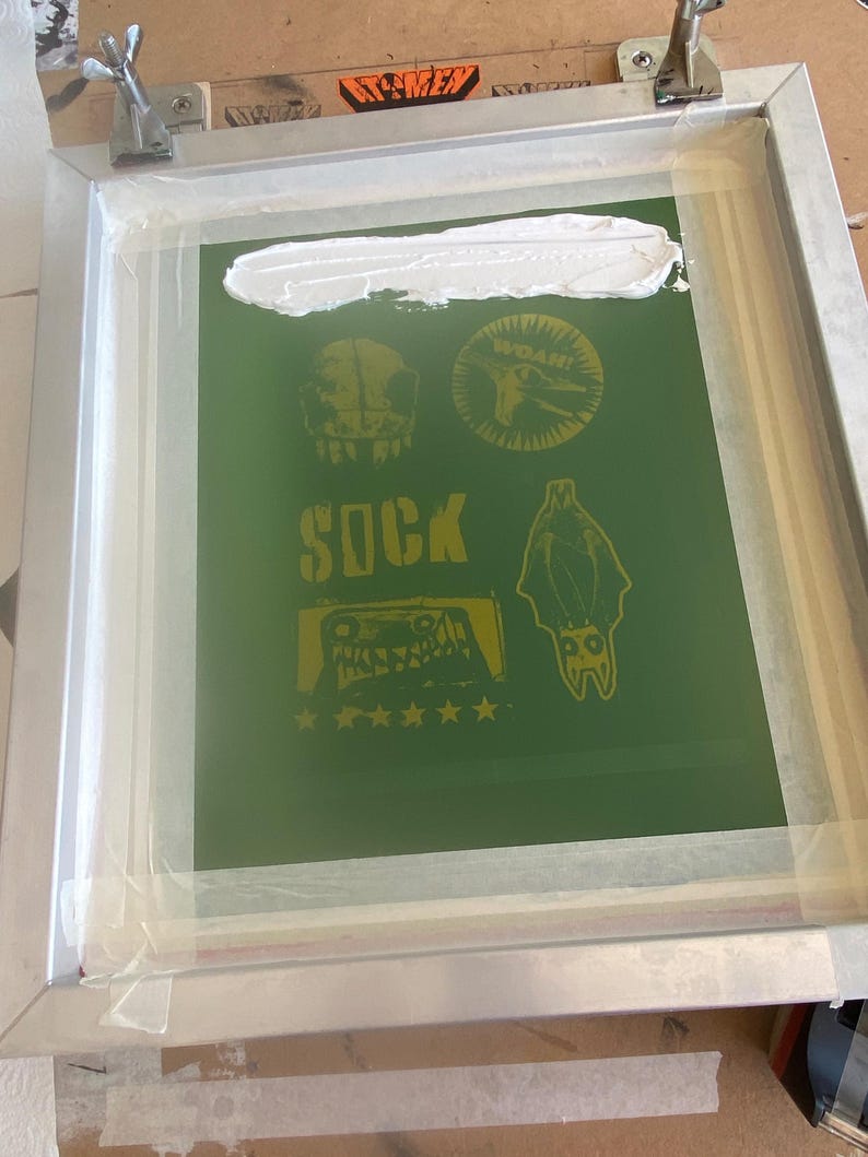Puede incluir: Un marco de serigraf&iacute;a con un fondo verde que presenta dise&ntilde;os gr&aacute;ficos amarillos, incluyendo una calavera, un murci&eacute;lago y la palabra "SOCK". Tinta blanca se aplica en la parte superior del marco. El marco est&aacute; asegurado con abrazaderas met&aacute;licas.