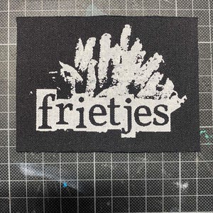 Puede incluir: Parche rectangular negro con la palabra "frietjes" en blanco, texto estilizado. Encima del texto hay un diseño abstracto blanco. El parche está sobre un fondo de cuadrícula.