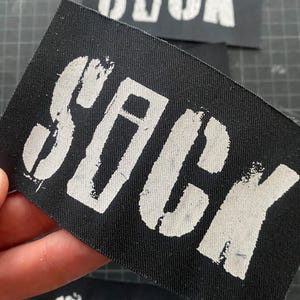 Könnte beinhalten: Schwarzer rechteckiger Aufnäher mit dem Wort "SICK" in weißen Buchstaben im Schablonenstil. Der Aufnäher besteht aus einem gewebten Stoff. Mehrere Aufnäher sind sichtbar, einer davon wird in einer Hand gehalten.