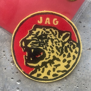 Könnte beinhalten: Runder Aufnäher mit rotem Rand und gelbem Jaguar-Design. Das Wort "JAG" ist oben in Weiß aufgedruckt. Ein kleiner gelber Aufkleber mit der Aufschrift "COOL STUFF" befindet sich im Hintergrund.