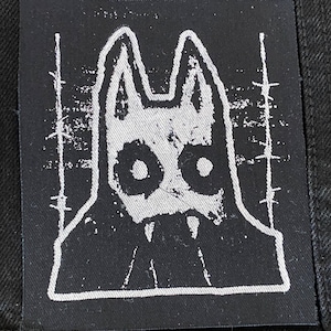 May include: Black square patch with a white illustration of a stylized wolf-like creature with large eyes and fangs. The design has a distressed, textured appearance, with vertical lines on the sides.