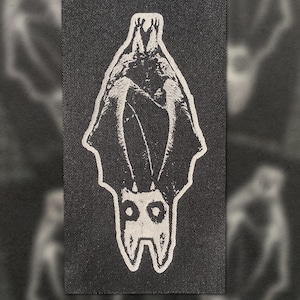 Könnte beinhalten: Schwarzer rechteckiger Aufnäher mit einem weißen Fledermaus-Design. Die Fledermaus ist kopfüber mit ausgebreiteten Flügeln dargestellt, darunter die Buchstaben "PO". Der Aufnäher hat eine strukturierte Oberfläche.