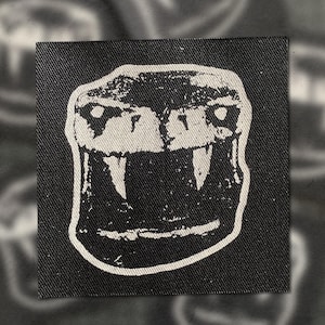 Peut inclure: Écusson noir et blanc carré avec une illustration stylisée d'une gueule de serpent ouverte avec des crocs. Le patch est fait d'un tissu texturé. Le design est une représentation graphique audacieuse.