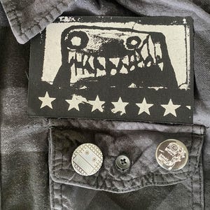 Puede incluir: Camisa a cuadros negros y grises con un parche rectangular con un gr&aacute;fico de monstruo y estrellas blancas. Dos alfileres redondos con dise&ntilde;os est&aacute;n en el bolsillo.