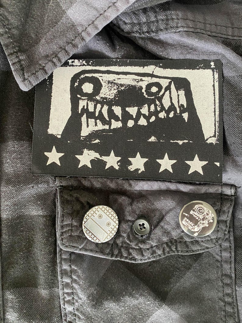 Puede incluir: Camisa a cuadros negros y grises con un parche rectangular con un gr&aacute;fico de monstruo y estrellas blancas. Dos alfileres redondos con dise&ntilde;os est&aacute;n en el bolsillo.