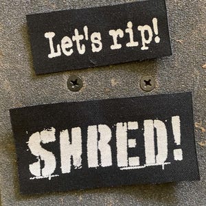 Könnte beinhalten: Zwei rechteckige schwarze Stoffaufnäher mit weißem gesticktem Text. Der obere Aufnäher zeigt den Schriftzug Let's rip! in einer Schreibmaschinenschrift, während der untere Aufnäher das Wort SHRED! in einer fetten, abgenutzten Blockschrift zeigt.