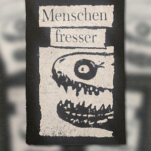 Pode incluir: Patch preto e branco com as palavras "Menschenfresser" e uma ilustração de monstro. O patch retangular tem uma borda preta e um fundo texturizado, adequado para roupas.